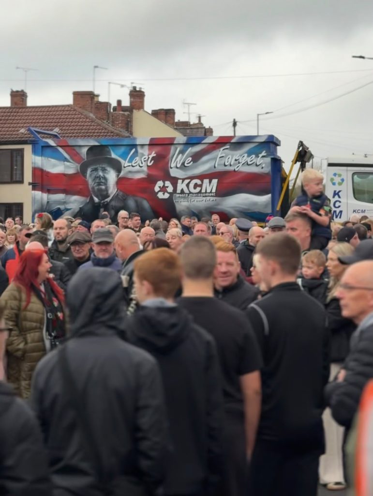 Crowd at Greasbrough’s 2025 Remembrance event beside a KCM vehicle displaying a “Lest We Forget” tribute.